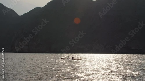 Tracking shot of silhouette of two people kayaking in the ocean next to towering limestone fjord mountains. Lens flare at top of frame, sun reflecting on the calm water in Khasab bay Musandam, Oman photo