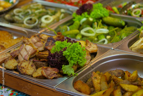 Various products are offered at a street food fair, event, festival. Fried potatoes and other foods on the counter next to large chunks of freshly cooked meat, garnished with bright green lettuce.