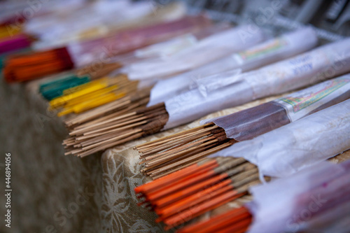 Multi-colored orange, yellow, red, green and brown incense sticks lie half visible in white paper wrappers on a counter covered in brown patterned cloth. Blurred background. Close-up.
