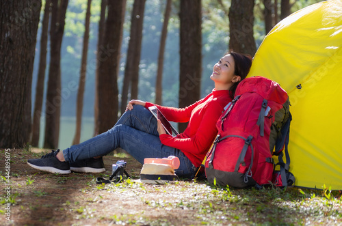 A woman leans against a yellow tent with a red backpack, She is use a tablet and enjoying nature in the midst of a beautiful pine forest beside lake, Pang Oung, Mae Hong Son, Thailand.