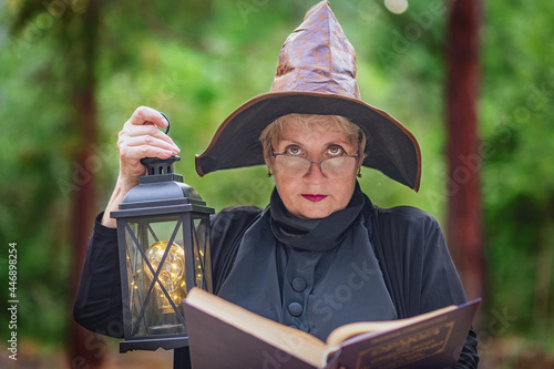 Fotografie An elderly witch in glasses in the woods, holding an open book, illuminating it with an antique lamp