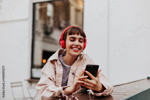 Positive short-haired girl in red headphones smiles outdoors. Brunette woman in beige jacket holding phone and listening to music outside.. photo