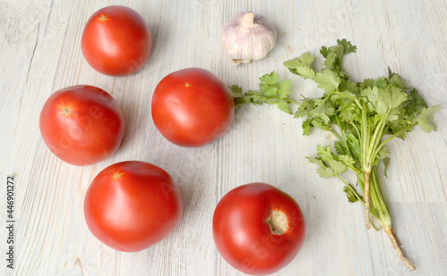 Five red large tomatoes are next to a bunch of greens and a head of garlic on a light background