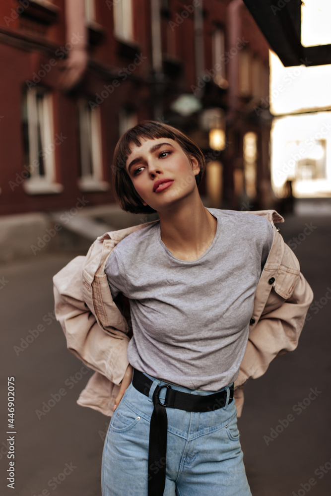 Fashionable lady with short hair posing in city. Trendy teen girl in beige denim jacket, jeans with black belt and grey t-short looking away outside..