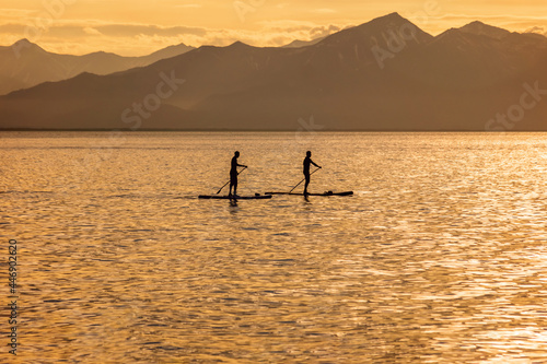Silhouettes of two men standing on sup board against the mountains on the ocean at sunset.