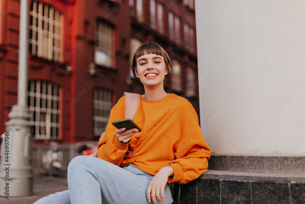 © Look! - Excited teen girl in orange sweatshirt leans on building and holds smartphone. Joyful woman in stylish hipster outfit smiles outside. © Look! - Excited teen girl in orange sweatshirt leans on building and holds smartphone. Joyful woman in stylish hipster outfit smiles outside.