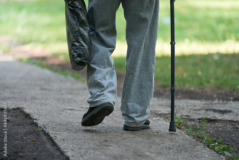 An elderly man slowly walks along the path leaning on a cane. In his hand he holds a plastic bag with the purchased products. An elderly man slowly walks along the path leaning on a cane. In his hand he holds a plastic bag with the purchased products.