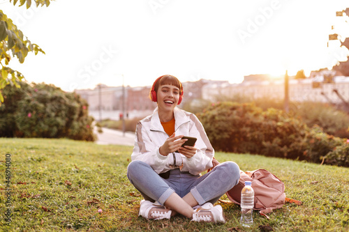 Cheerful short-haired girl sits on grass during sunset. Pretty woman in white jacket and jeans smiles, listens to music in headphones.
