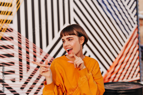 Good-humored lady with brunette hair pointing her fingers on striped backdrop. Modern girl in orange clothes smiling outside..
