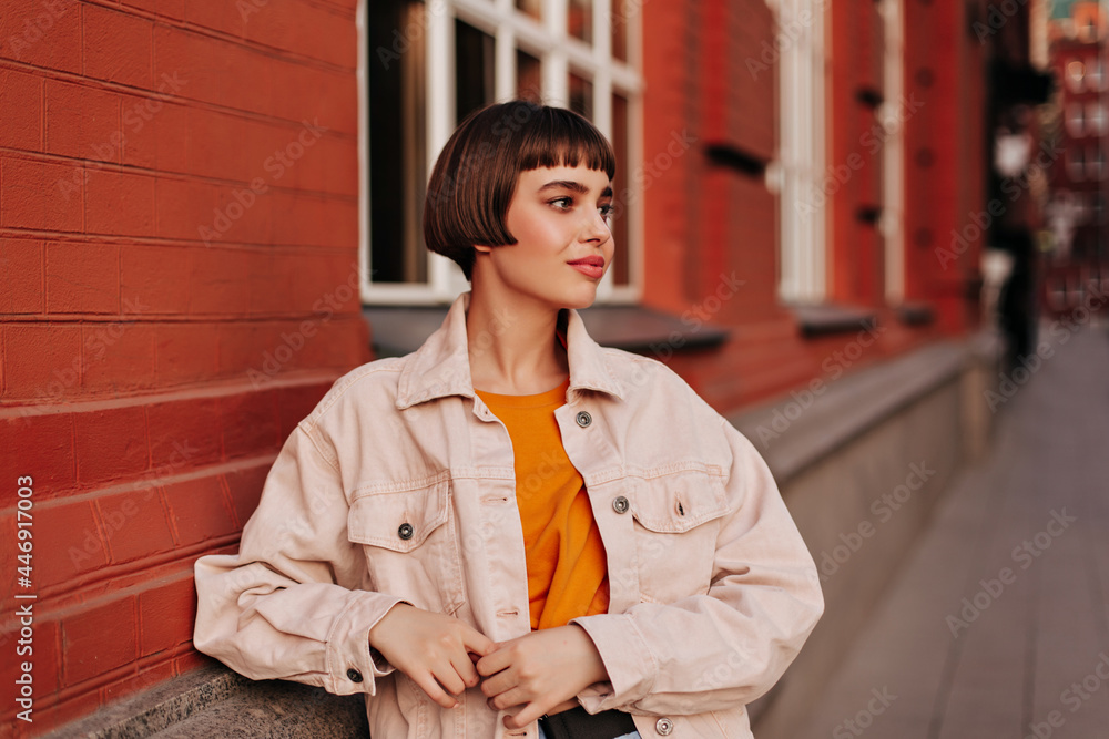 Cool woman in pink denim jacket and orange sweatshirt poses outside near brick building. Attractive girl walks outdoors. - obrazy, fototapety, plakaty 