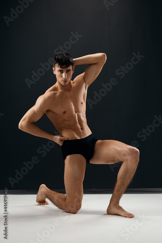 a sporty man with a naked body in black panties on a dark background is on his knee