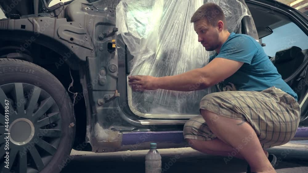 A painter worker who seals the holes of a car and other body elements with paint tape so as not to fill it with paint during painting in a car repair shop. Pasting with a film. A painter worker who seals the holes of a car and other body elements with paint tape so as not to fill it with paint during painting in a car repair shop. Pasting with a film.