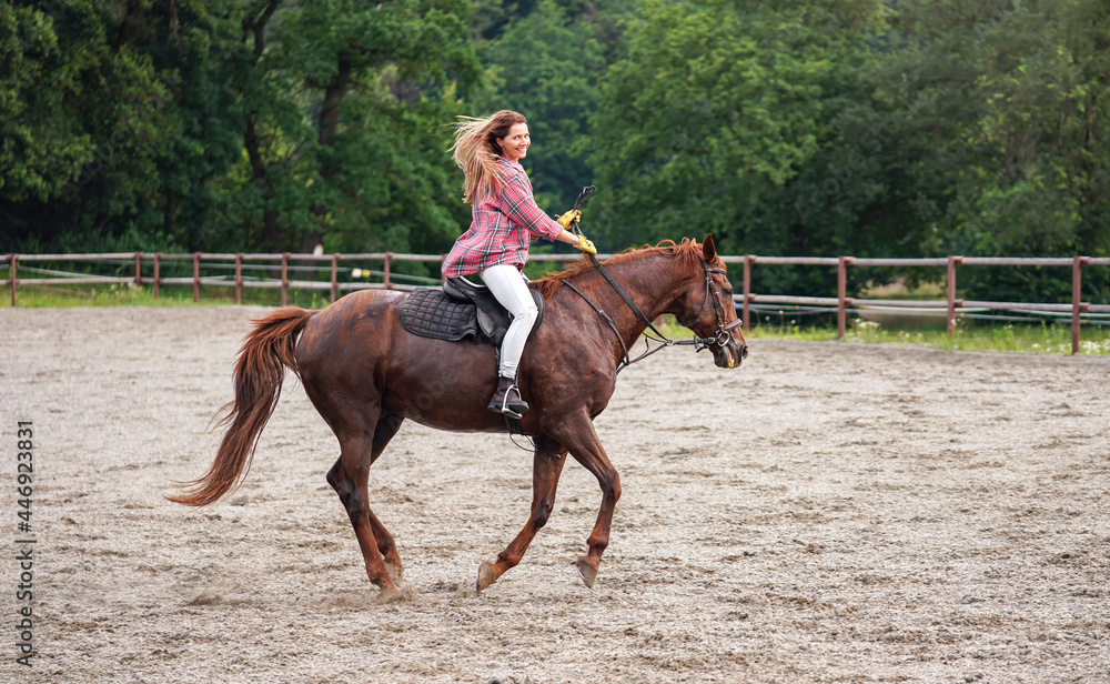 Young woman in shirt riding on brown horse, smiling, hair moving in air because of speed, blurred trees background