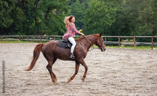 Young woman in shirt riding on brown horse, smiling, hair moving in air because of speed, blurred trees background © Lubo Ivanko