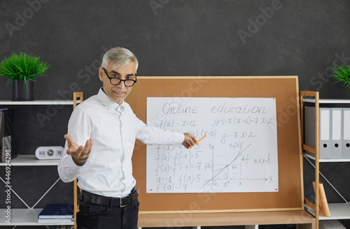 Teacher having online class. Portrait of intelligent smart mature man in glasses standing in front of classroom board, pointing at it, looking at camera and asking question during virtual math lesson