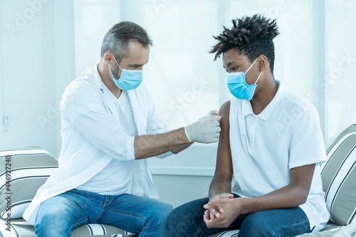 doctor wearing a mask holds a syringe for the COVID-19 vaccine to an African American teenager at a vaccination station. 