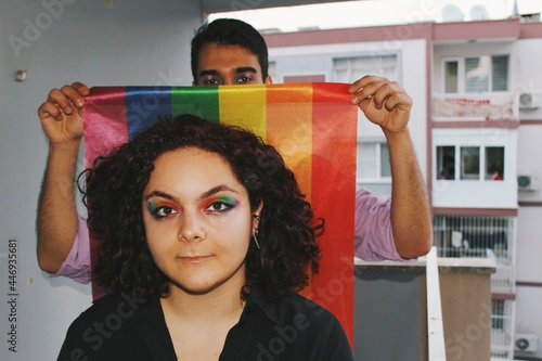 portrait of lgbtq young woman with queer friend holding rainbow pride flag in home balcony