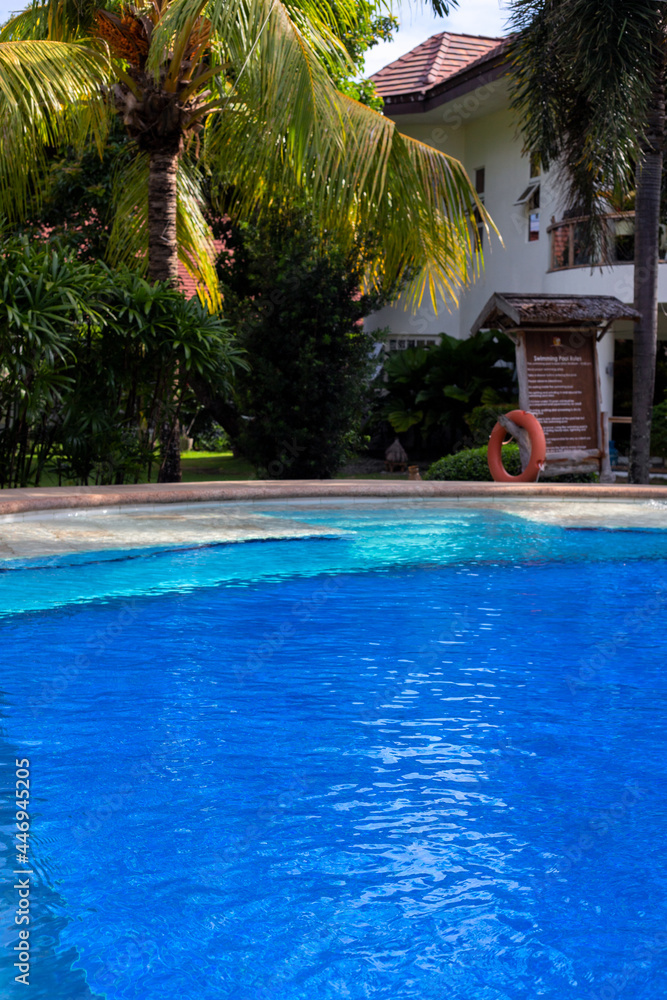 Fototapeta premium Swimming pool with palm tree and hotel building. Fresh water pool banner template. Empty amenities of hotel or resort. Pandemic travel ban. Sunny swimming pool with clean water ripples. Pool service