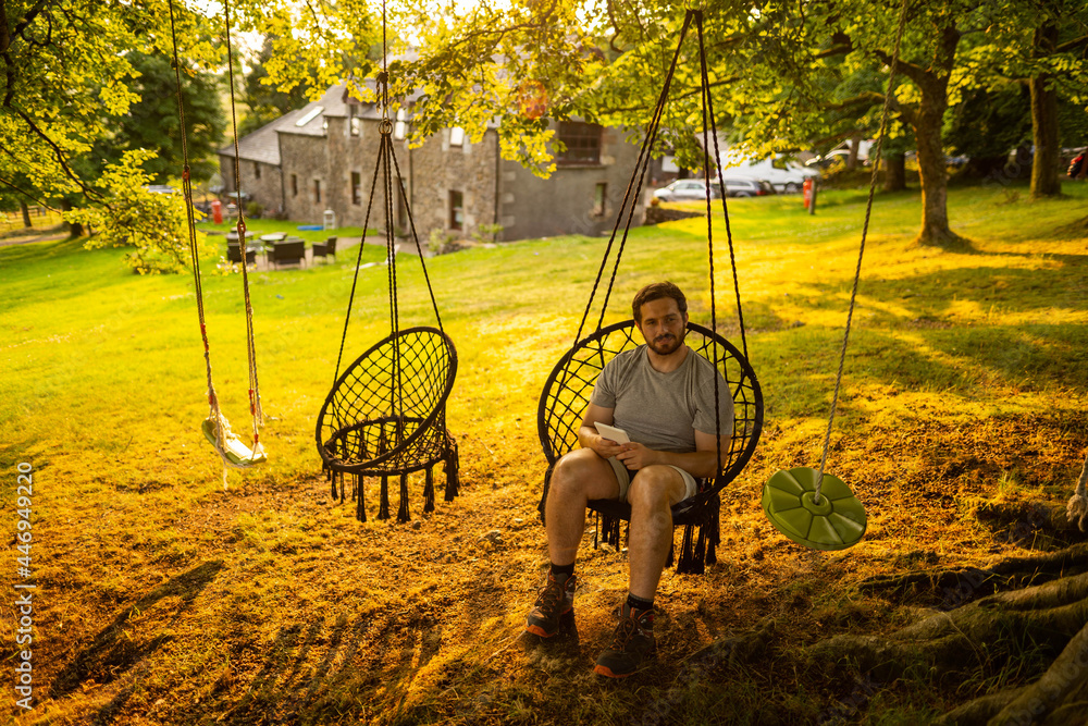 Photo of a young and attractive guy sitting on a hammock relaxed reading an electronic book. It is a summer day in the countryside of Scotland