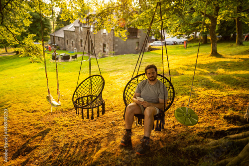 Obraz na płótnie Photo of a young and attractive guy sitting on a hammock relaxed reading an electronic book. It is a summer day in the countryside of Scotland w salonie