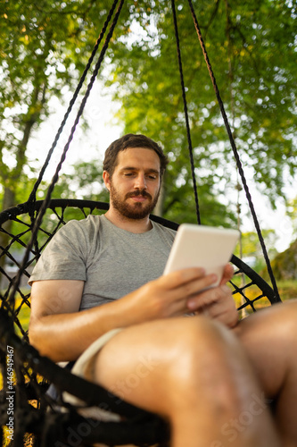 Photo of a young and attractive guy sitting on a hammock relaxed reading an electronic book. It is a summer day in the countryside of Scotland