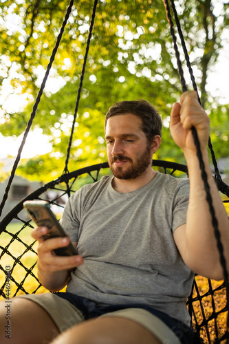 Photo of a guy and attractive bearded guy relaxing on a hammock and checking his phone while enjoying a summer day in the countryside in Scotland
