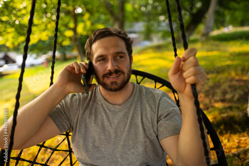 Photo of a guy and attractive bearded guy relaxing on a hammock and checking his phone while enjoying a summer day in the countryside in Scotland