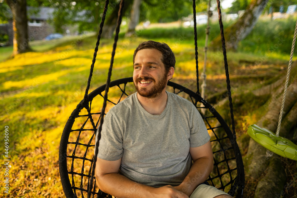 Photo of a attractive young guy relaxing on a hammock in the middle of nature. It is a summer day in the forest and he has a happy smile