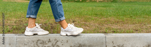 A woman in jeans and sneakers walks along the curb. Legs in sneakers close-up, banner.