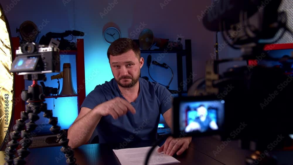 Cacasian man blogger with scar in his hand in blue T-shirt is broadcasting in video studio with cameras and professional lighting. In hands of sheets of paper with text news. Dolly shot Cacasian man blogger with scar in his hand in blue T-shirt is broadcasting in video studio with cameras and professional lighting. In hands of sheets of paper with text news. Dolly shot