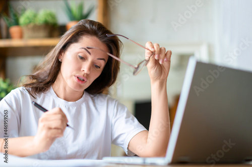 A woman works with papers at home in front of a laptop monitor. She is sitting with glasses and checking papers.