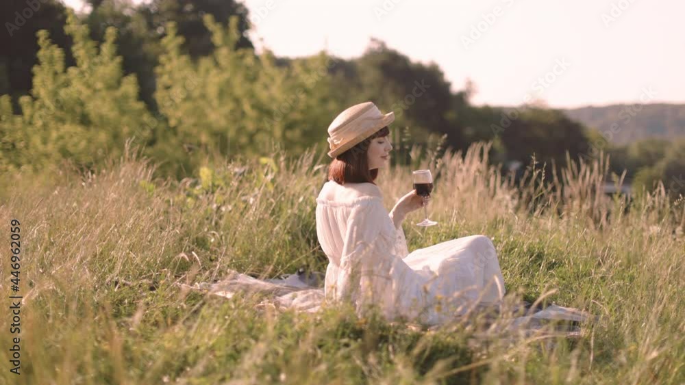 Pretty 30-aged European lady, wearing white dress and stylish straw hat, sitting on the blanket outdoors in the field, drinking red wine and enjoying wonderful nature ladscape Pretty 30-aged European lady, wearing white dress and stylish straw hat, sitting on the blanket outdoors in the field, drinking red wine and enjoying wonderful nature ladscape