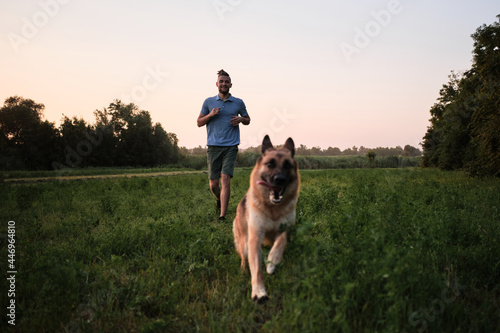 Young handsome Caucasian man with beard and dreadlocks is walking with dog on warm summer evening in park. Jogging with German Shepherd in field. Sports and recreation with dog.