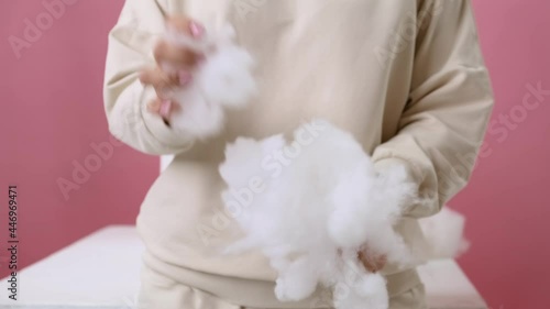 Medium shot. A girl in a vanilla-colored hoodie on a pink background tears up a voluminous artificial filler