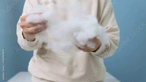 Medium shot. A girl in a vanilla-colored hoodie on a blue background tears up a voluminous artificial filler of white color