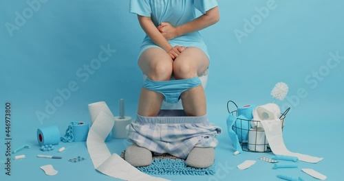 Unknown person poses on toilet bowl touches stomach suffers from abdominal cramps upleasant feelings because of digestion or diarrhea wears slippers pants on legs poses in restroom against blue wall photo