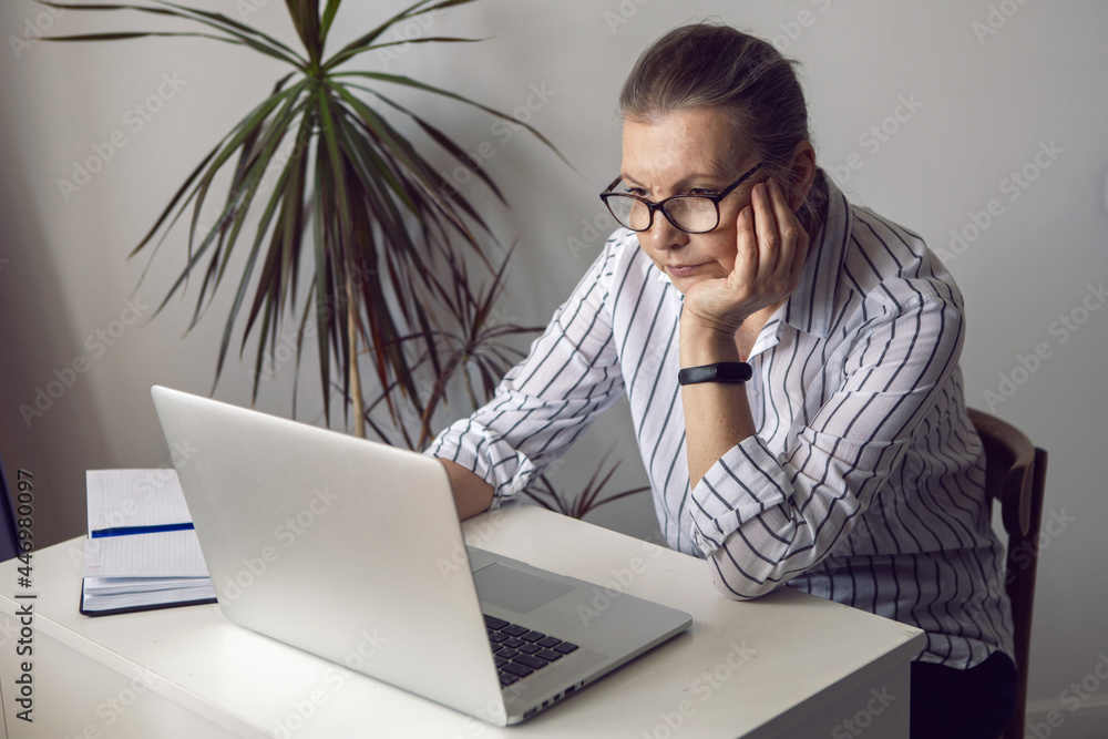 woman in a white striped shirt aged sits at a computer at home with glasses woman in a white striped shirt aged sits at a computer at home with glasses