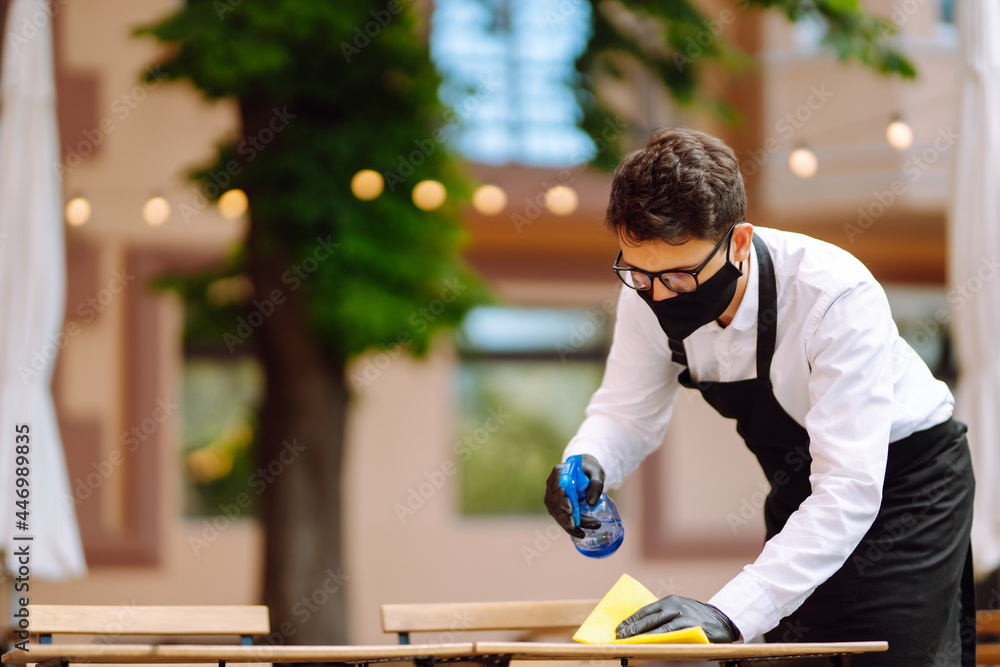 Disinfecting to prevent COVID-19. Waiter in protective face mask and gloves cleaning and disinfecting restaurant table for next customer. Disinfecting to prevent COVID-19. Waiter in protective face mask and gloves cleaning and disinfecting restaurant table for next customer.