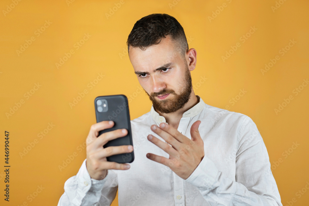 A young man in a white business shirt on a yellow background holds a phone and looks at it and is angry because he was written bad news, the shares of his company are depreciating A young man in a white business shirt on a yellow background holds a phone and looks at it and is angry because he was written bad news, the shares of his company are depreciating