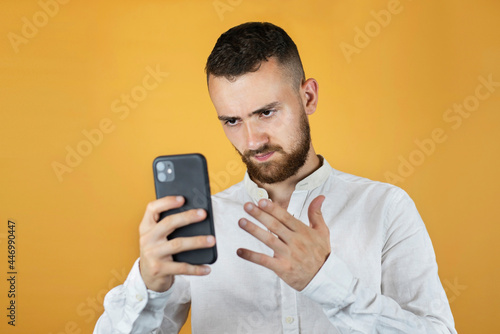 A young man in a white business shirt on a yellow background holds a phone and looks at it and is angry because he was written bad news, the shares of his company are depreciating