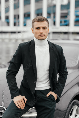 A handsome young man in a black suit is standing near a sports car. A man with a watch on his hands in a dark business suit