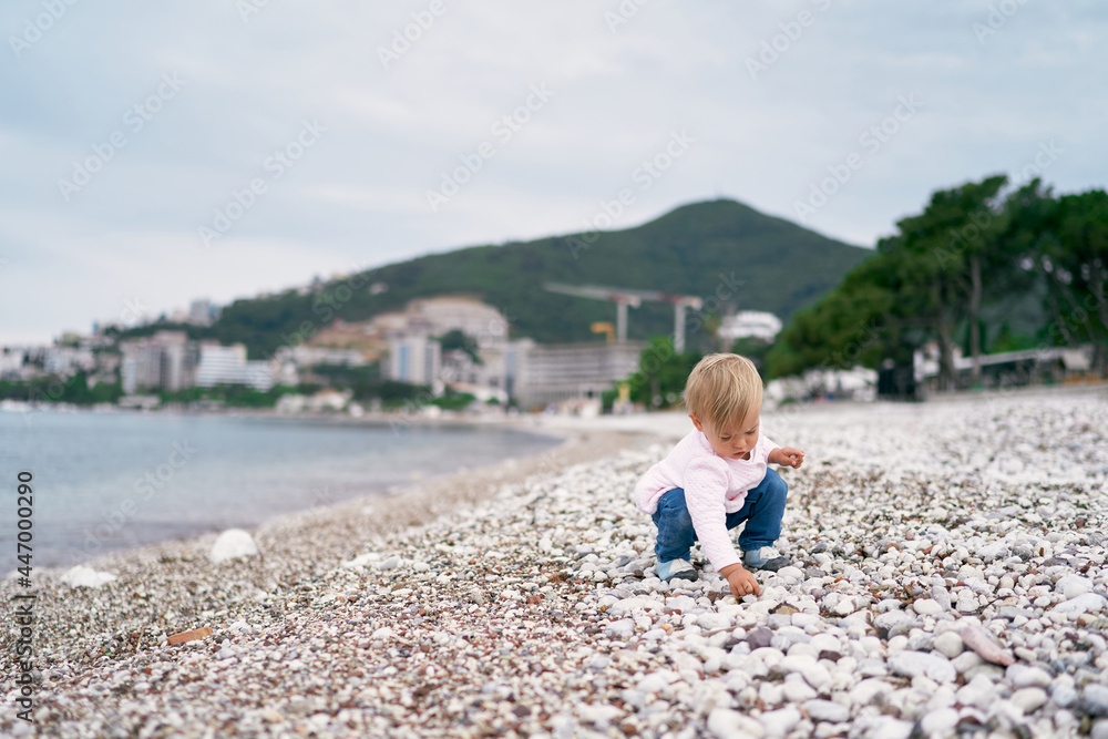 Little girl squatting on a pebble beach and holding a pebble in her hand