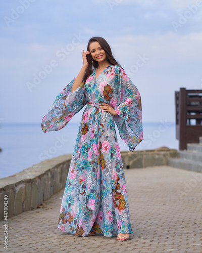 Full length outdoor portrait of young elegant tall woman in colorful blue dress with floral print walking at sea ambankment on a sunny day