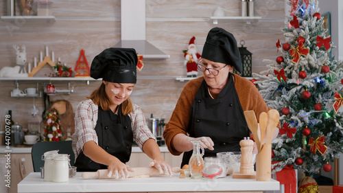 Grandma coffee grandchild to prepare homemade gingerbread dessert in culinary kitchen during traditional christmas holidays. Child with apron making dough using rolling pin enjoying christmastime photo