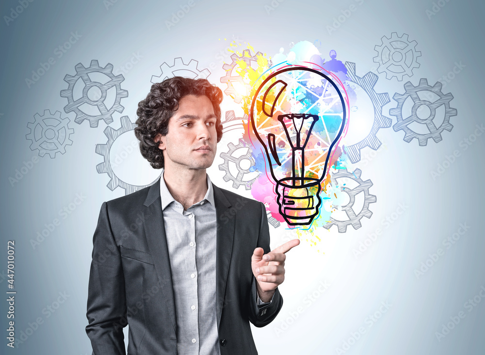 Hispanic handsome businessman in suit pointing out by finger and pondering about new ideas. Blue wall with colorful business idea light bulb and cog wheels sketches. Creativity and brainstorming Hispanic handsome businessman in suit pointing out by finger and pondering about new ideas. Blue wall with colorful business idea light bulb and cog wheels sketches. Creativity and brainstorming