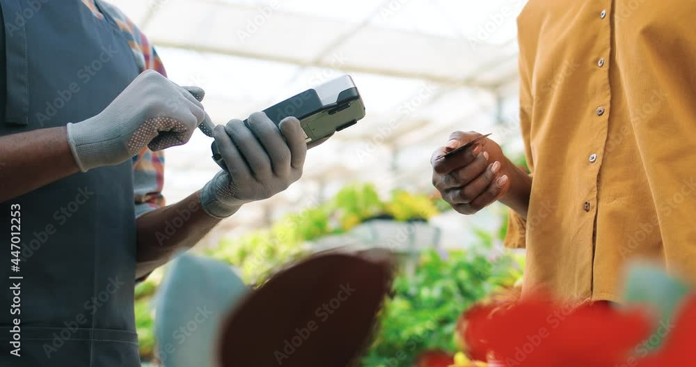 Close up of African American female hands using credit card paying for purchase using payment terminal in flower shop. Client buying plants making payment in store. Shopping in garden center, business Close up of African American female hands using credit card paying for purchase using payment terminal in flower shop. Client buying plants making payment in store. Shopping in garden center, business