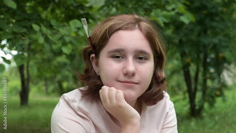 Portrait of a teenage girl Looking into the camera with an art brush behind her ear in a park on a Summer day. A Young Sad Thinking Curious Child Looks at the camera Close-up in the open air. The Face Portrait of a teenage girl Looking into the camera with an art brush behind her ear in a park on a Summer day. A Young Sad Thinking Curious Child Looks at the camera Close-up in the open air. The Face