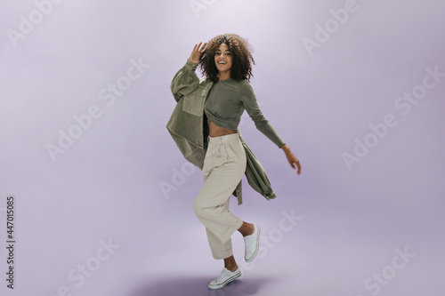 Fashionable cool girl with brunette wavy hairstyle in olive top, trendy jacket, white pants and modern sneakers smiling.