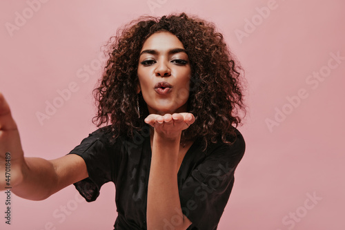 Photo of women with curly short hairstyle and earrings in black fashionable polka dot clothes making selfie and blowing kiss.