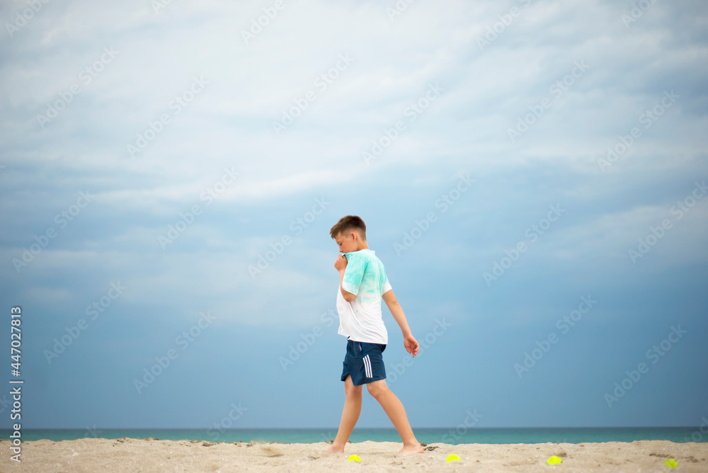 Boy training on a sandy beach. Tennis balls are spread out on the coast. Rest between exercises.